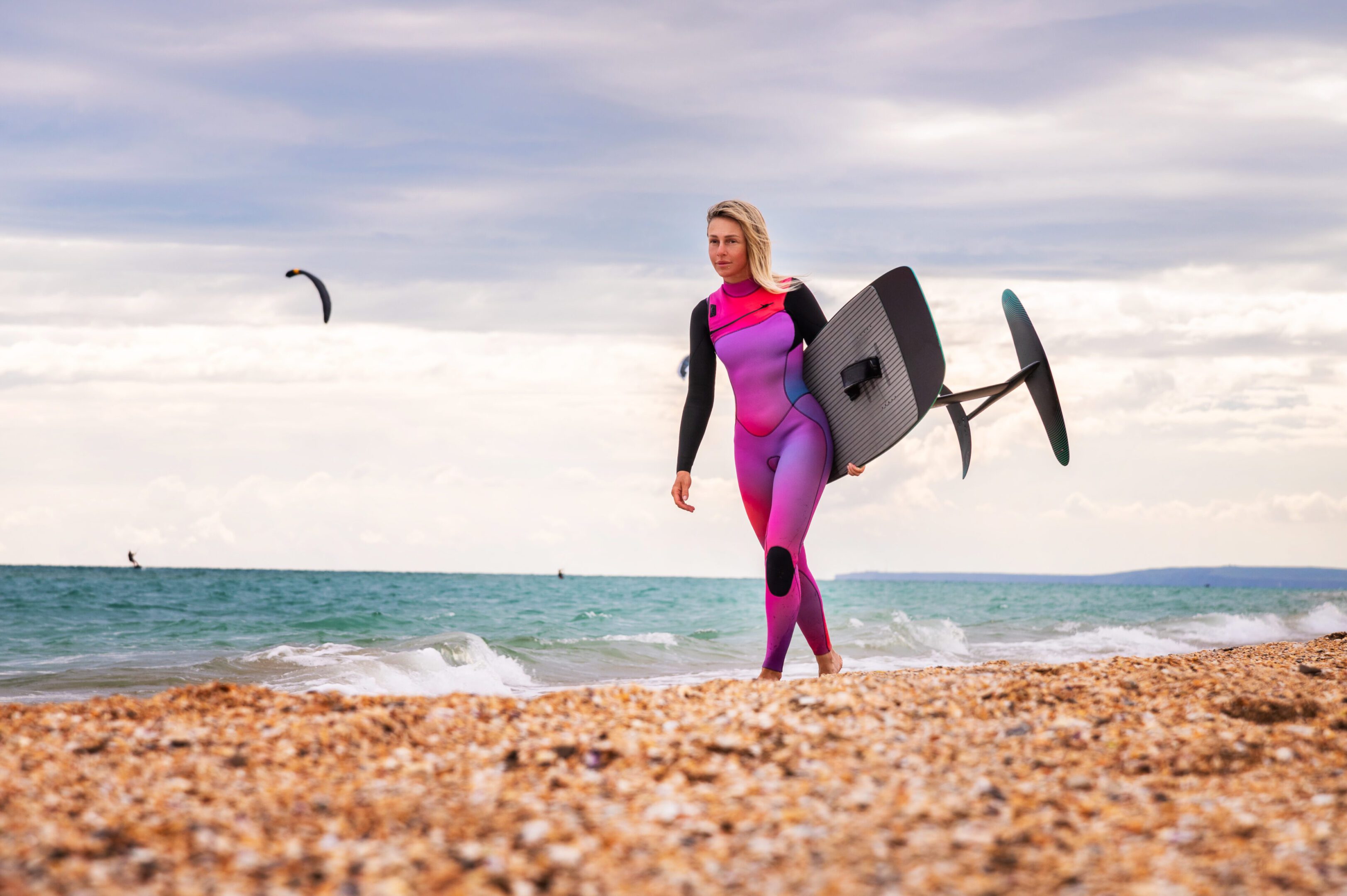 A woman in purple wetsuit carrying a surfboard.